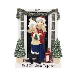 Op de afbeelding: Een gepersonaliseerde kerstversiering met een stel dat voor een deur met een krans staat. De versiering is wit met een rode en groene krans, een zwarte deur en een rode lantaarn. Het stel draagt winterkleding en houdt elkaar vast. De versiering heeft de tekst "2025" en "Eerste Kerst samen".