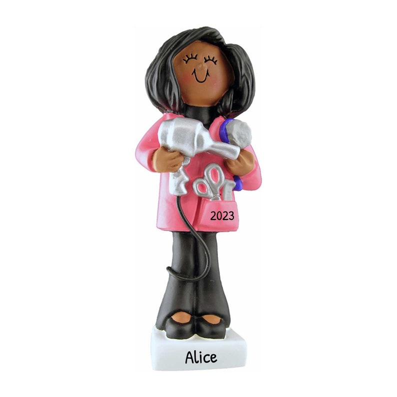 May include: A figurine of a hairstylist holding a silver hairdryer and a blue hairbrush. The figure wears a pink coat over a black top and pants. The figurine has black hair and is standing on a white base with the name "Alice" and the year "2023" printed on it.