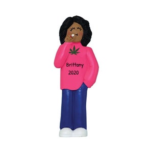 May include: A pink Christmas ornament featuring a cartoon woman with black hair, wearing a pink sweater with a green marijuana leaf and the text "Brittany 2020".