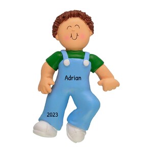 Op de afbeelding: Een gepersonaliseerde kerstversiering met een jongen met bruin haar die een blauwe overall en een groen shirt draagt. De versiering is versierd met de naam "Adrian" en het jaar "2023".