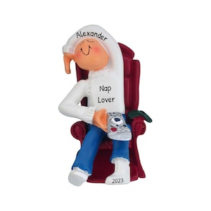 May include: A white personalized Christmas ornament depicting a person in a red armchair wearing a white robe with the text "Nap Lover" and the name "Alexander". The ornament also includes the year "2023".
