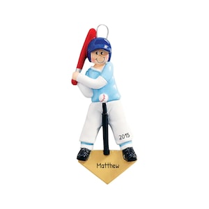 May include: A baseball-themed ornament featuring a figure in a blue helmet and uniform, holding a red bat. The ornament includes a baseball on a tee, a base with the name "Matthew", and the year "2015".