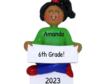 May include: A personalized Christmas ornament featuring a girl with brown skin and black hair holding a sign that says "6th Grade!" The sign is white with black text. The girl is wearing a green shirt with the name "Amanda" written on it, blue pants, and red shoes. The ornament also has the year "2023" written on it.