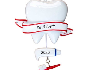 May include: White tooth-shaped Christmas ornament with a red ribbon that says "Dr. Robert" and a white tube of toothpaste with the year "2020" on it. A red toothbrush with white bristles is hanging from the tube of toothpaste.