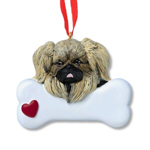 May include: A Christmas ornament featuring a Pekingese dog with brown and black fur, peeking over a white bone with a red heart. The ornament is made of ceramic and has a gold string for hanging.