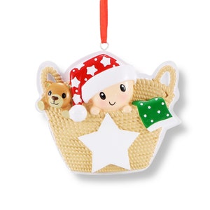 May include: A personalised Christmas bauble featuring a baby in a basket with a teddy bear and a star. The baby is wearing a red and white Santa hat and a green blanket with white polka dots. The bauble is made of white ceramic and has a gold ribbon for hanging.