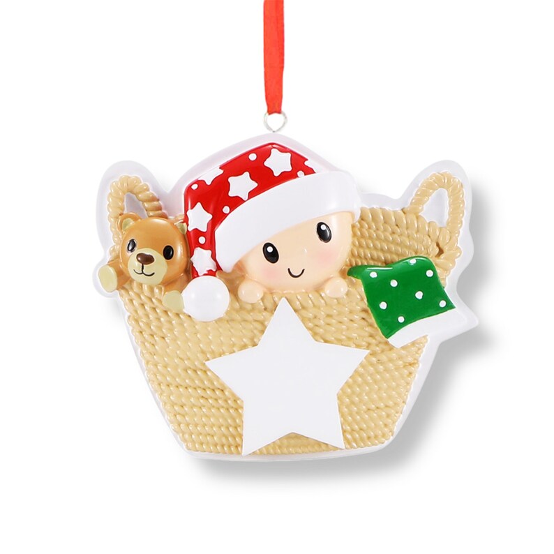 May include: A personalised Christmas bauble featuring a baby in a basket with a teddy bear and a star. The baby is wearing a red and white Santa hat and a green blanket with white polka dots. The bauble is made of white ceramic and has a gold ribbon for hanging.