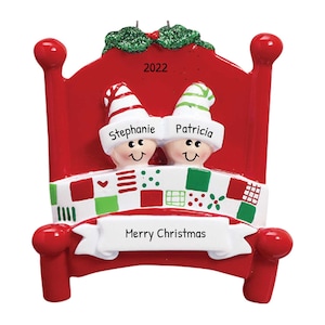 May include: Red and white Christmas ornament shaped like a bed with two elves in it. The elves are wearing green and red hats and have the names "Stephanie" and "Patricia" written on them. The bed has a patchwork quilt and a banner that says "Merry Christmas". The year "2022" is written on the headboard.