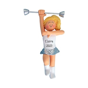 May include: A personalized ornament of a girl twirling a baton. She is wearing a white leotard with a silver skirt and white boots. The ornament is decorated with the name "Claire" and the year "2023".