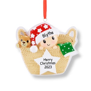 May include: A personalised Christmas bauble featuring a baby in a basket with a teddy bear. The baby is wearing a red and white Santa hat with the name "Blythe" written on it. The bauble also includes the text "Merry Christmas 2023".
