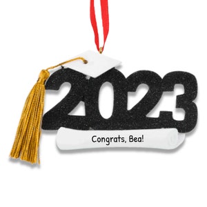 以下が含まれることがあります： 2023年と金のタッセルが付いた黒い卒業帽子のオーナメント。白い巻物に「おめでとう、ベア！」という文字が書かれており、下に付けられています。