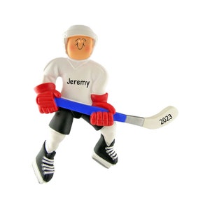 May include: White hockey player Christmas ornament with a blue and silver hockey stick. The player is wearing a white jersey with the name "Jeremy" on the front. The hockey stick has the year "2023" on it.