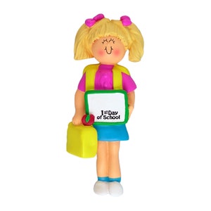 May include: A girl doll ornament with blonde hair, wearing a pink shirt, blue skirt, and a yellow backpack. She is holding a green chalkboard with the text "1st Day of School".