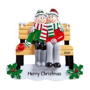 May include: A Christmas ornament featuring a couple sitting on a bench with a red cardinal. The ornament is decorated with snow, holly, and the text "Merry Christmas". The couple is wearing winter hats and scarves, and the woman's hat says "Katherine" and the man's hat says "Benjamin". The bench has the year "2020" on it.