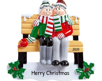 May include: A Christmas ornament featuring a couple sitting on a bench with a red cardinal. The ornament is decorated with snow, holly, and the text "Merry Christmas". The couple is wearing winter hats and scarves, and the woman's hat says "Katherine" and the man's hat says "Benjamin". The bench has the year "2020" on it.