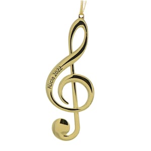 May include: A gold-colored treble clef ornament with black accents. The ornament has the text "Alicia 2022" engraved on it. The ornament is suspended by a gold-colored ribbon.