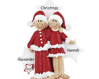 May include: A personalised Christmas bauble featuring a couple in red pyjamas with white trim, standing on white stars. The bauble includes the names "Alexander", "Hannah", and "Christmas" and the year "2021".