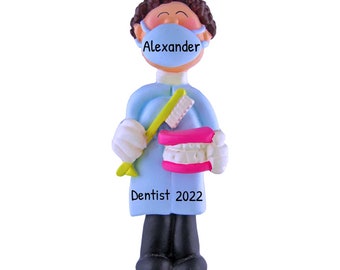 May include: A blue and white personalised ornament of a dentist wearing a face mask and holding a toothbrush and a set of teeth. The ornament says "Alexander Dentist 2022".