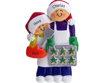 May include: Personalized Christmas ornament featuring a couple wearing white aprons and Santa hats. The woman is holding a spatula and the man is holding a baking sheet with star-shaped cookies. The ornament is decorated with green glitter and the year 2023. The names "Nova" and "Charles" are written on the hats.
