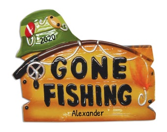 Könnte beinhalten: Ein dekoratives Ornament in Form eines Holzschildes mit der Aufschrift GONE FISHING in schwarzen Buchstaben. Ein grüner Hut mit der Jahreszahl 2020 liegt obenauf, dazu eine Angelrute mit Schnur und Haken. Der Name Alexander ist schwarz aufgedruckt.