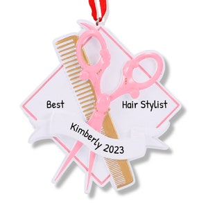 Op de afbeelding: Witte en gouden kerstversiering met een roze schaar en een kam. De versiering heeft de tekst "Best Hair Stylist Kimberly 2023" erop.