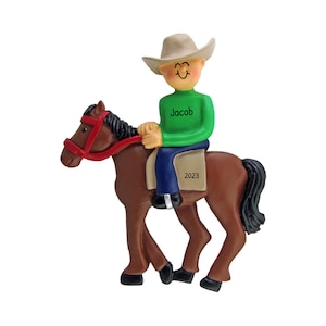 May include: A brown horse ornament with a green shirt wearing cowboy riding on top. The shirt has the name "Jacob" written on it. The ornament is dated "2023".