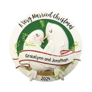 May include: A white Christmas ornament with two white doves kissing. The ornament is round and has a green background with a red ribbon that says "A Very Married Christmas" and "Gracelynn and Jonathan". The year 2024 is printed on the bottom of the ornament.