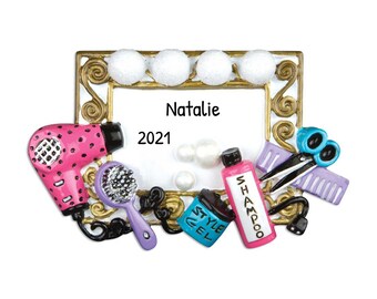 May include: A gold-colored picture frame with white bulbs around the top. The frame has a white background with the name "Natalie" and the year "2021" written in black. The frame is surrounded by various hair styling tools, including a pink hair dryer, a purple hairbrush, a blue bottle of styling gel, a pink bottle of shampoo, a pair of silver scissors, and a purple comb.