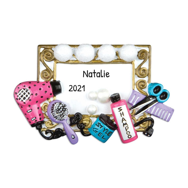 May include: A gold-colored picture frame with white bulbs around the top. The frame has a white background with the name "Natalie" and the year "2021" written in black. The frame is surrounded by various hair styling tools, including a pink hair dryer, a purple hairbrush, a blue bottle of styling gel, a pink bottle of shampoo, a pair of silver scissors, and a purple comb.