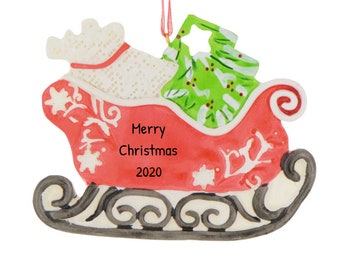 Puede incluir: Adorno navideño rojo y blanco con forma de trineo con una bolsa blanca de regalos y un árbol de Navidad verde en su interior. El trineo tiene un borde negro y blanco y el texto "Feliz Navidad 2020" en el lateral.