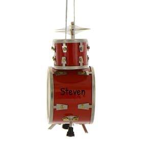 May include: A red miniature drum kit ornament with silver-coloured hardware and the name "Steven" printed on the bass drum. The ornament includes a cymbal and is suspended by a silver string.