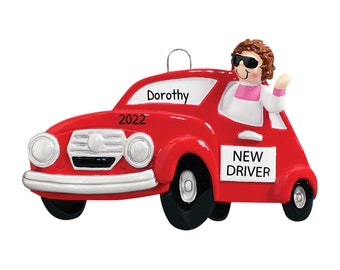 May include: Red car Christmas ornament with a woman driver wearing sunglasses and a pink shirt. The car has a white sign that says "NEW DRIVER" and the year "2022" on the front. The car has a white sign on the back that says "Dorothy".
