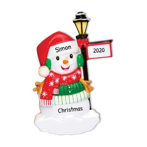 May include: A snowman Christmas ornament with a red Santa hat, green scarf, and a sign that says "2020". The snowman is holding a streetlight and the base of the ornament says "Christmas". The snowman's hat has the name "Simon" written on it.