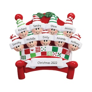 May include: A red and white Christmas ornament shaped like a bed with eight elf heads peeking out from under the covers. The ornament is decorated with a patchwork quilt design and the text "Christmas 2022".