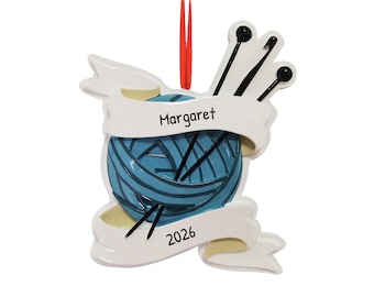 May include: A white ceramic Christmas ornament with a blue yarn ball, knitting needles, and a banner that reads "Margaret 2026".