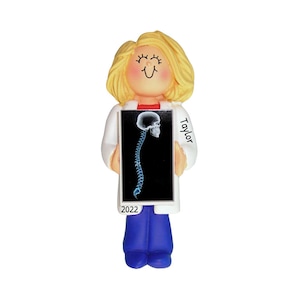 May include: A white-coated figurine of a person with blonde hair holding a black and white x-ray image of a spine. The figurine is wearing blue pants and has a smiling face. The x-ray image has the text "Taylor" written on it. The year "2022" is written on the bottom of the figurine.