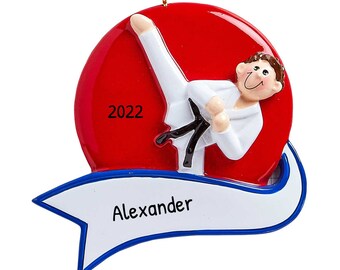 May include: A red and white personalized Christmas ornament featuring a cartoon figure in a white karate uniform with a black belt, kicking in the air. The ornament has a blue and white banner with the name "Alexander" and the year "2022" printed on the red background.