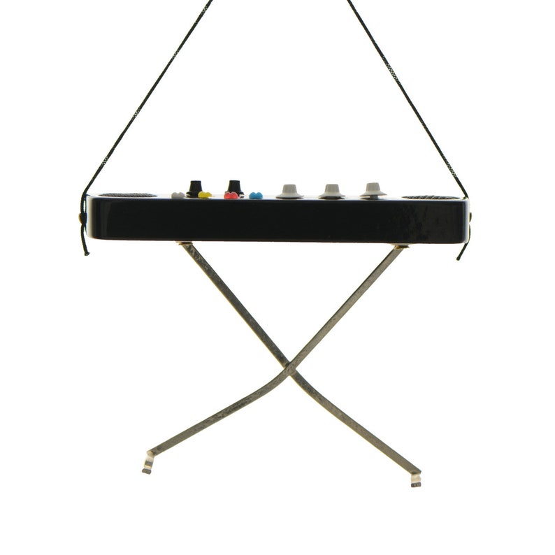 May include: A miniature black synthesizer with colorful knobs and buttons, suspended by a black string. It rests on a silver, X-shaped stand. The synthesizer has a sleek, glossy finish and a minimalist design, perfect for music enthusiasts.