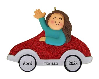 May include: Red glitter car ornament with a girl waving from the driver's seat. The car has white wheels with the names "April" and "Marissa" and the year "2024" written on them.