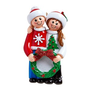 Puede incluir: Un adorno navide&ntilde;o con una pareja sosteniendo una corona con un marco de fotos. El hombre lleva un su&eacute;ter rojo con un dise&ntilde;o de copo de nieve y la mujer lleva un su&eacute;ter blanco con un dise&ntilde;o de &aacute;rbol de Navidad. Ambos llevan gorros de Pap&aacute; Noel.