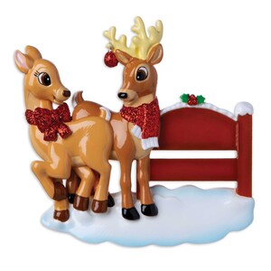 Puede incluir: Adorno navide&ntilde;o con dos renos. Uno lleva una bufanda roja con lentejuelas, el otro un lazo rojo con lentejuelas. Se encuentran frente a una valla roja con acebo y nieve, una encantadora decoraci&oacute;n navide&ntilde;a.