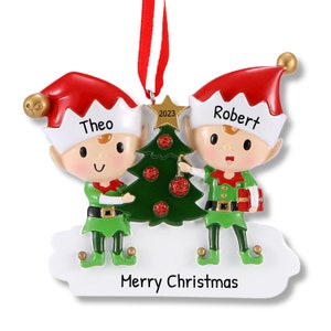Op de afbeelding: Gepersonaliseerde kerstversiering met twee elven die een kerstboom versieren. De elven dragen groene en rode outfits en hebben de namen Theo en Robert op hun hoeden. De versiering bevat ook het jaar 2023 en de tekst "Prettige Kerst".