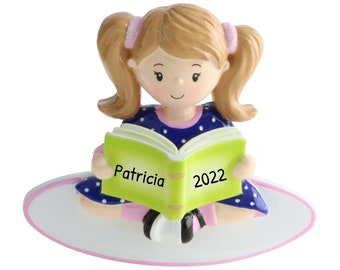May include: A personalized Christmas ornament featuring a girl with pigtails sitting on a white oval base, reading a green book with the name "Patricia" and the year "2022" written on it.