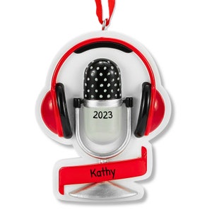 May include: A red and silver Christmas ornament featuring a microphone with headphones and a banner that says "Kathy". The microphone has a black and white polka dot design and the year "2023" is printed on the front.