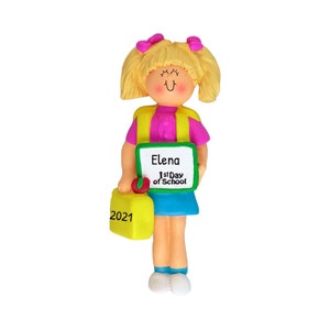 May include: A personalized ornament featuring a girl with blonde hair and a pink shirt holding a green chalkboard with the text "Elena 1st Day of School" and a yellow lunchbox with the year "2021".