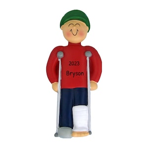 May include: A figurine of a person with a cast on their leg, using crutches for support. The person is wearing a red shirt, blue pants, and a green hat. The text "2023 Bryson" is printed on the shirt.
