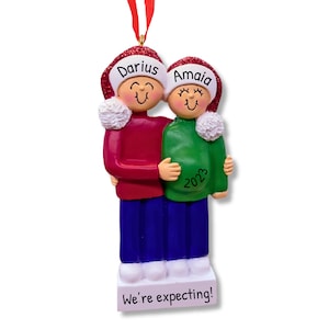 Op de afbeelding: Gepersonaliseerde kerstversiering met een stel in kerstmanmutsen, waarbij de vrouw een babybuik heeft. De versiering heeft de tekst "Darius" en "Amaia" op de mutsen en "2023" op het shirt van de vrouw. De basis van de versiering heeft de tekst "We're expecting!"