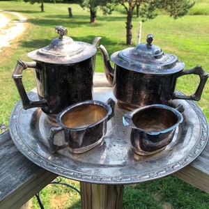 May include: A vintage silver tea set, including a teapot, creamer, sugar bowl, and serving tray. The set has ornate detailing and is displayed outdoors on a wooden surface with a green background.