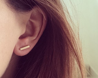 May include: A close-up of a person's ear with a gold bar earring. The earring is simple and minimalist, and it is a popular style of jewelry.