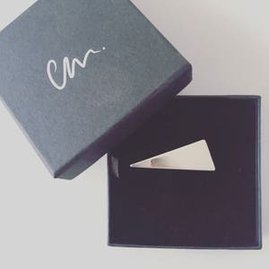May include: A silver triangle shaped ring in a black velvet box. The box has the initials "CM" written on the top.